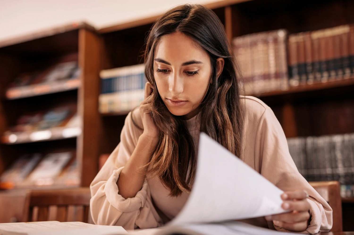 Estudiante concentrada revisando documentos en una biblioteca, rodeada de estanterías con libros.